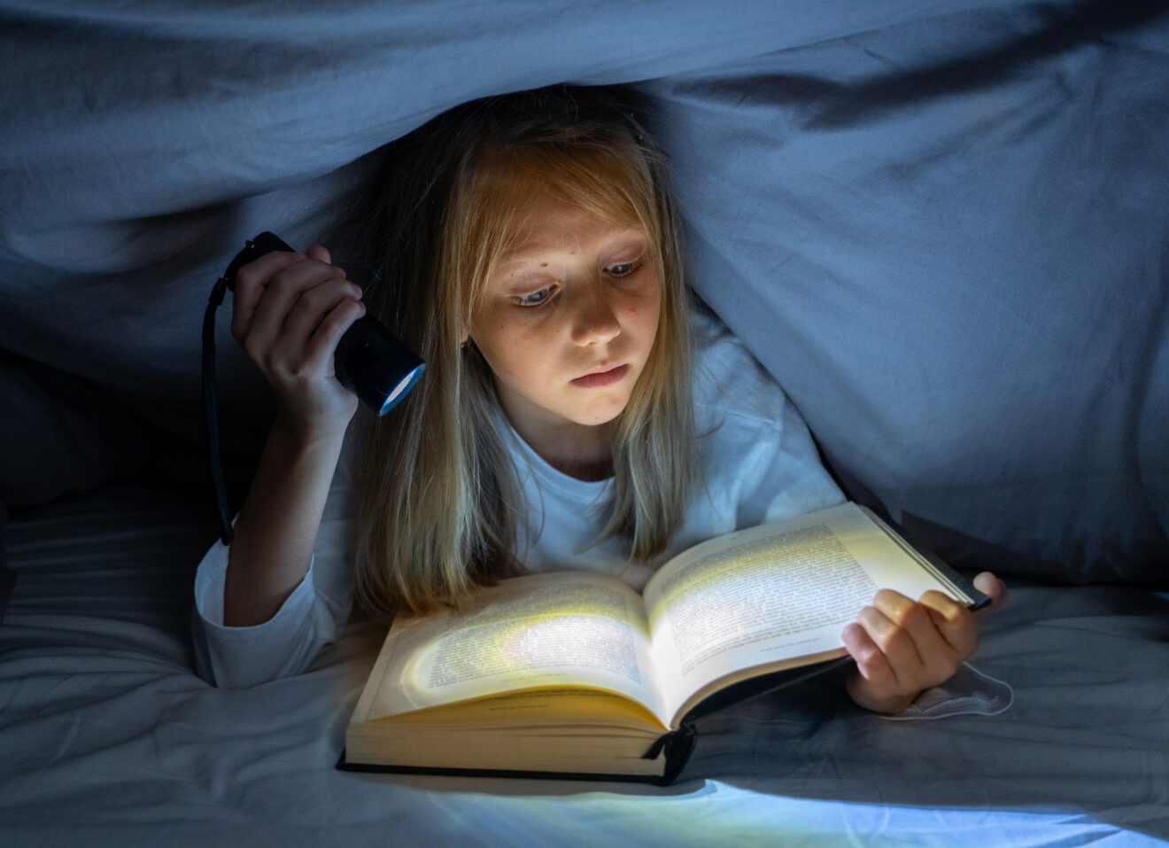 Scared little girl reading scary book under bed cover holding a